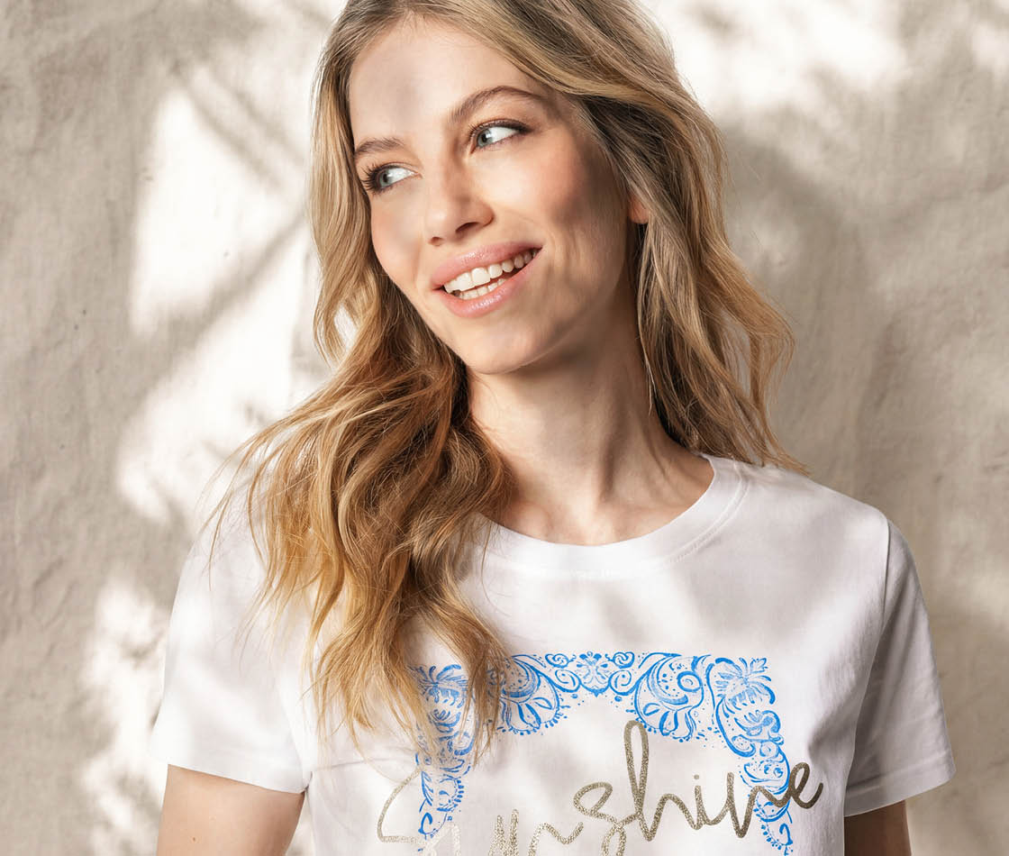 Dame im Mykonos-Moments-Look mit gemustertem Sommer-Outfit vor mediterraner Kulisse