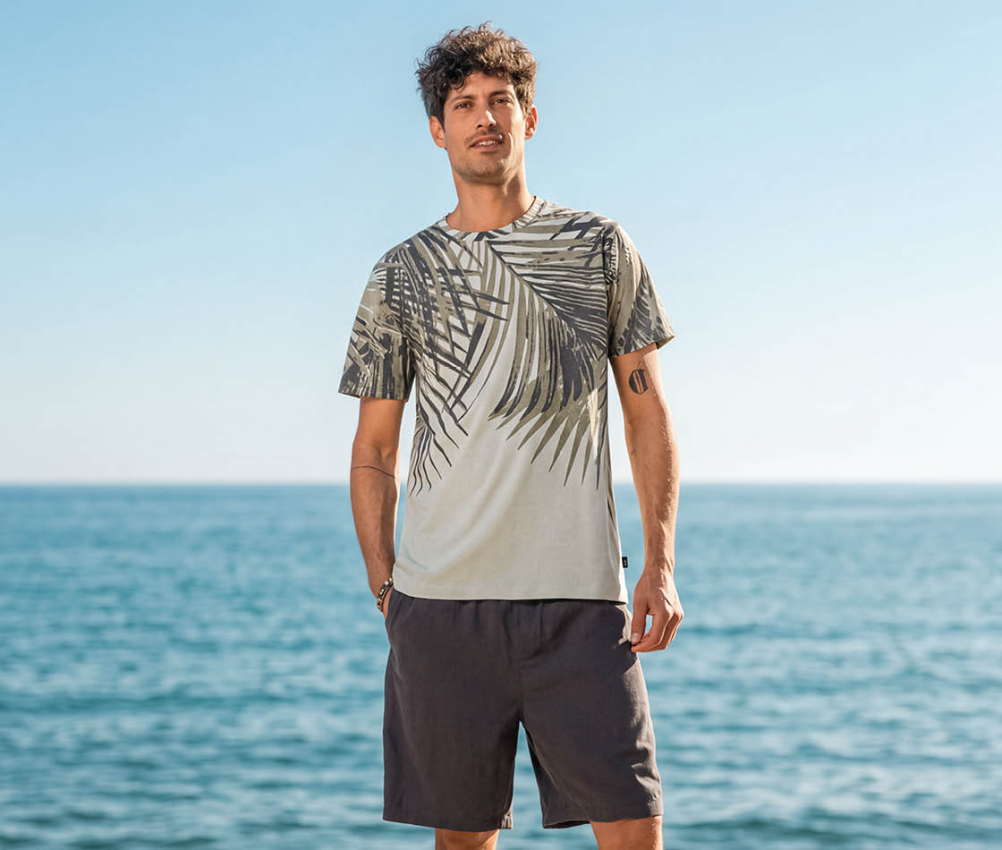 Mann am Meer im hellen T-Shirt mit dunklem Palmen-Print und dunkelgrauen Shorts